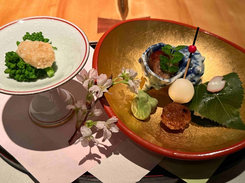 金地の盆に並ぶ前菜。子持ちヤリイカの旨煮、百合根クリームチーズ、合鴨つくね串、桜葉に包んだ寿司。手前に菜花のとろろ掛けと桜の枝