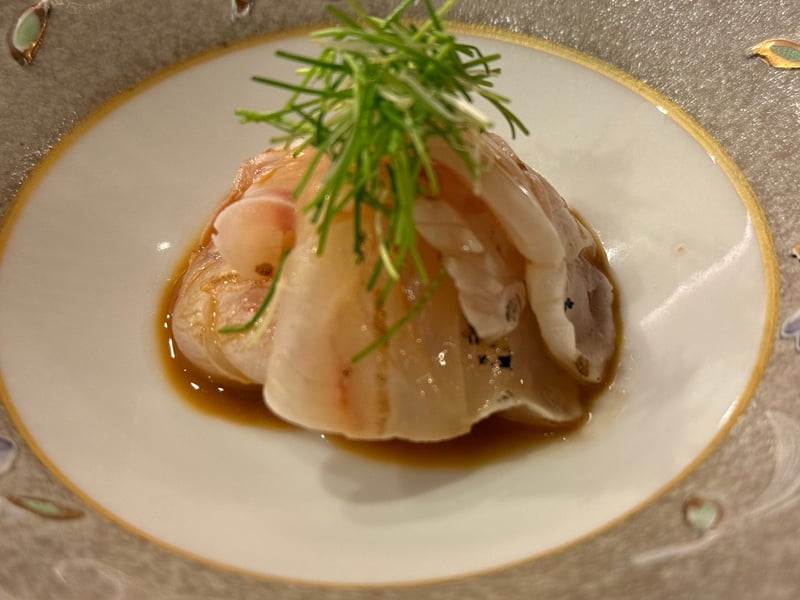 金縁の白い皿に重ねたクエの薄造り。細い芽ねぎをのせ、下に醤油のたまり