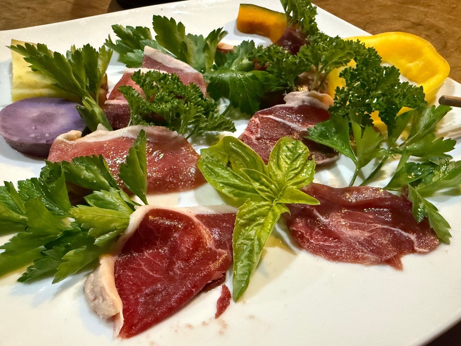 白い角皿に放射状に並べた生の獣肉の薄切り。パセリやバジルの葉、紫のじゃがいも、黄色いビーツ、かぼちゃが添えてある