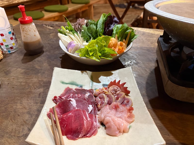 鍋の用意。皿に鹿と猪の赤身の薄切りと鶏、鉢にほうきたけや葉野菜、奥のコンロに鍋がかかっている