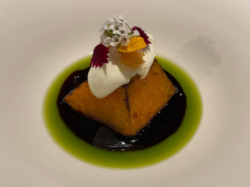 白い皿に、焼いたかぼちゃを黒いソースと緑のオイルで囲み、上にクリームと花