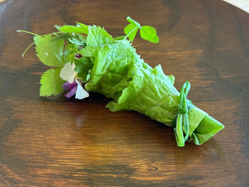木の皿の上、レタスの葉で束ねた野草とハーブのブーケ。紫と白の花がのぞく