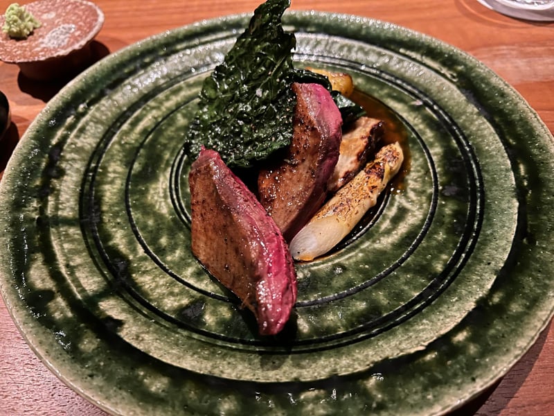 織部の緑の皿に、中心が赤く残るピジョンの胸肉二切れ。焼いたホワイトアスパラガスと素揚げの青菜