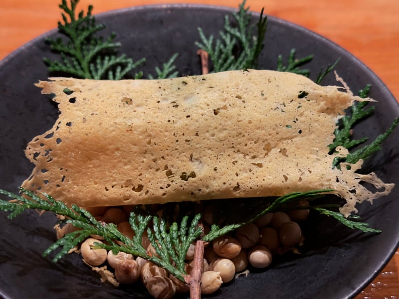 黒い皿に炒った豆とヒバの枝を敷き、その上に草を散らしたチーズのチュイールを一枚かぶせた一皿
