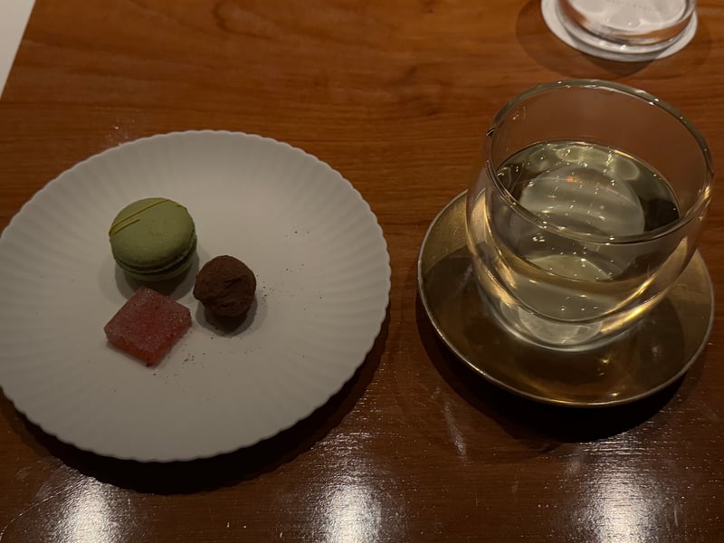 白い皿に抹茶色のマカロン、チョコレートの粒、桃色の四角い菓子。横にグラスの飲みもの