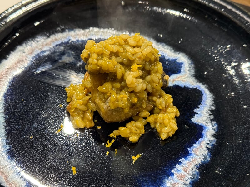 藍の流れる黒い皿に、鮑をのせた緑がかったリゾット。黄色い柑橘の皮が散らしてある