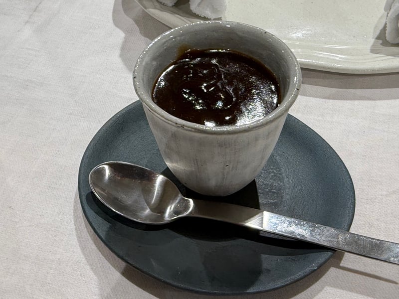 小さな白い器に入った濃い茶色のとろりとしたもの。黒い受け皿にスプーンが添えてある