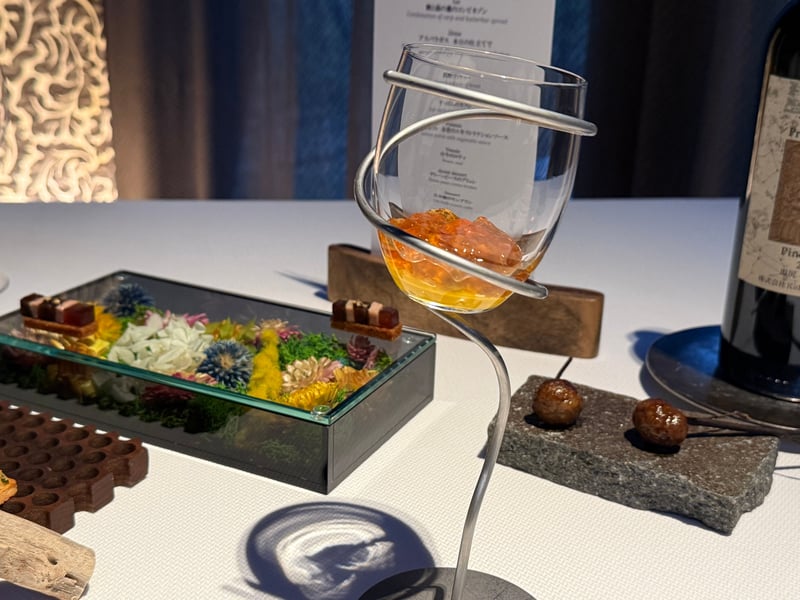 ドライフラワーを敷いたガラスのケースにのった小さな菓子と、螺旋の金具に支えられた人参色のムースのグラス、石板の上の茶色いソーセージ2粒