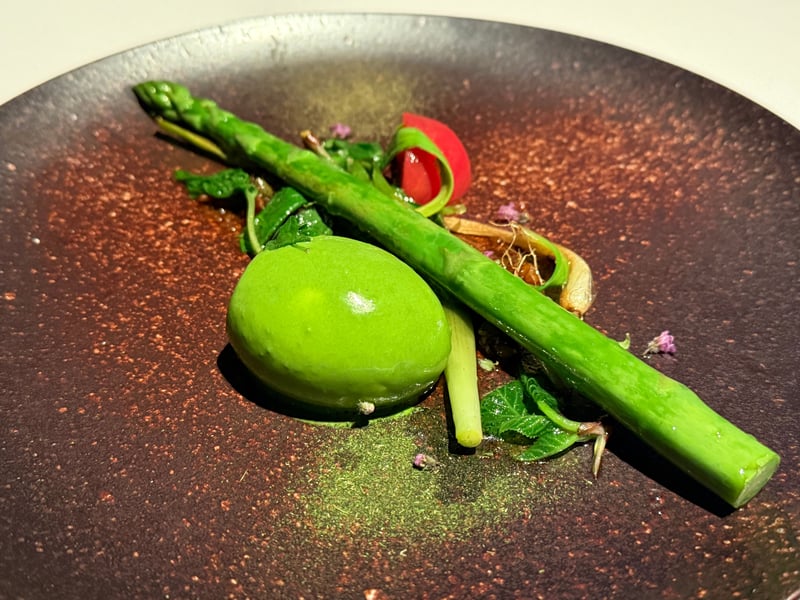茶色い斑の皿に、丸ごとのアスパラガス1本と緑のクリームのクネル、青菜と赤い野菜、紫の小花