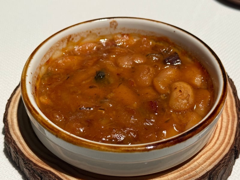 木の円盤にのせた白いココット。白い豆とトマト色の煮汁が縁まで入っている