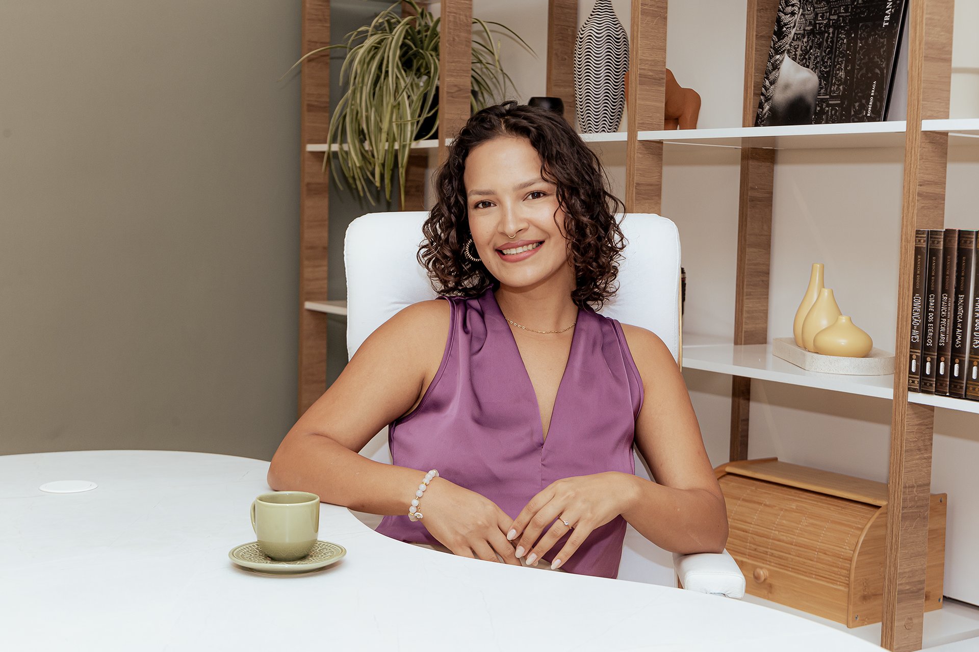 Mulher branca de cabelos enrolados na altura do pescoço sentada na cadeira de escritório branca ela está sorrindo vestida de blusinha roxa