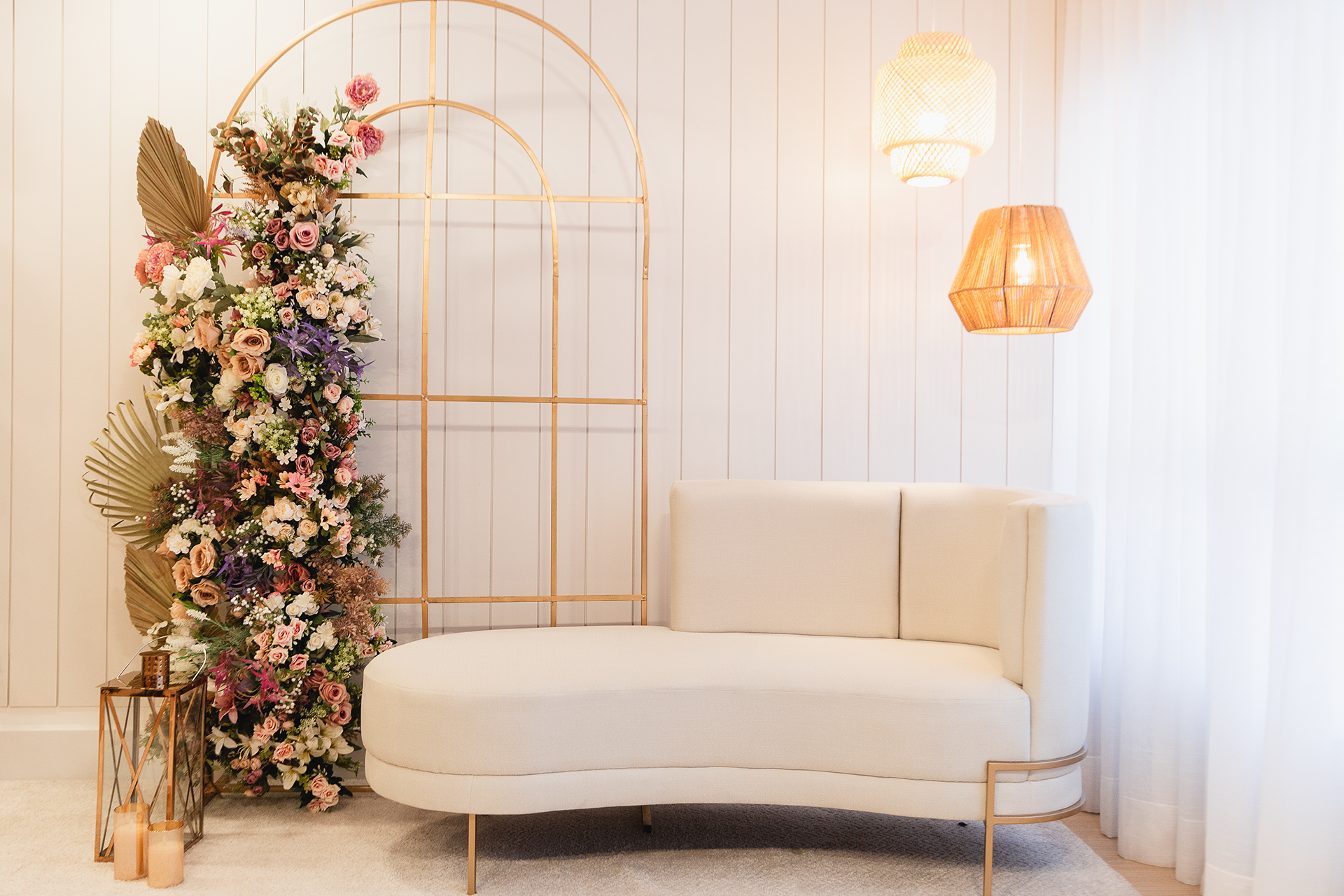 Nosso cenário para o dia das mães: Um floral em um aramado dourado na esquerda, um sofá off-white redonto, pendentes de madeira, e uma cortina branca