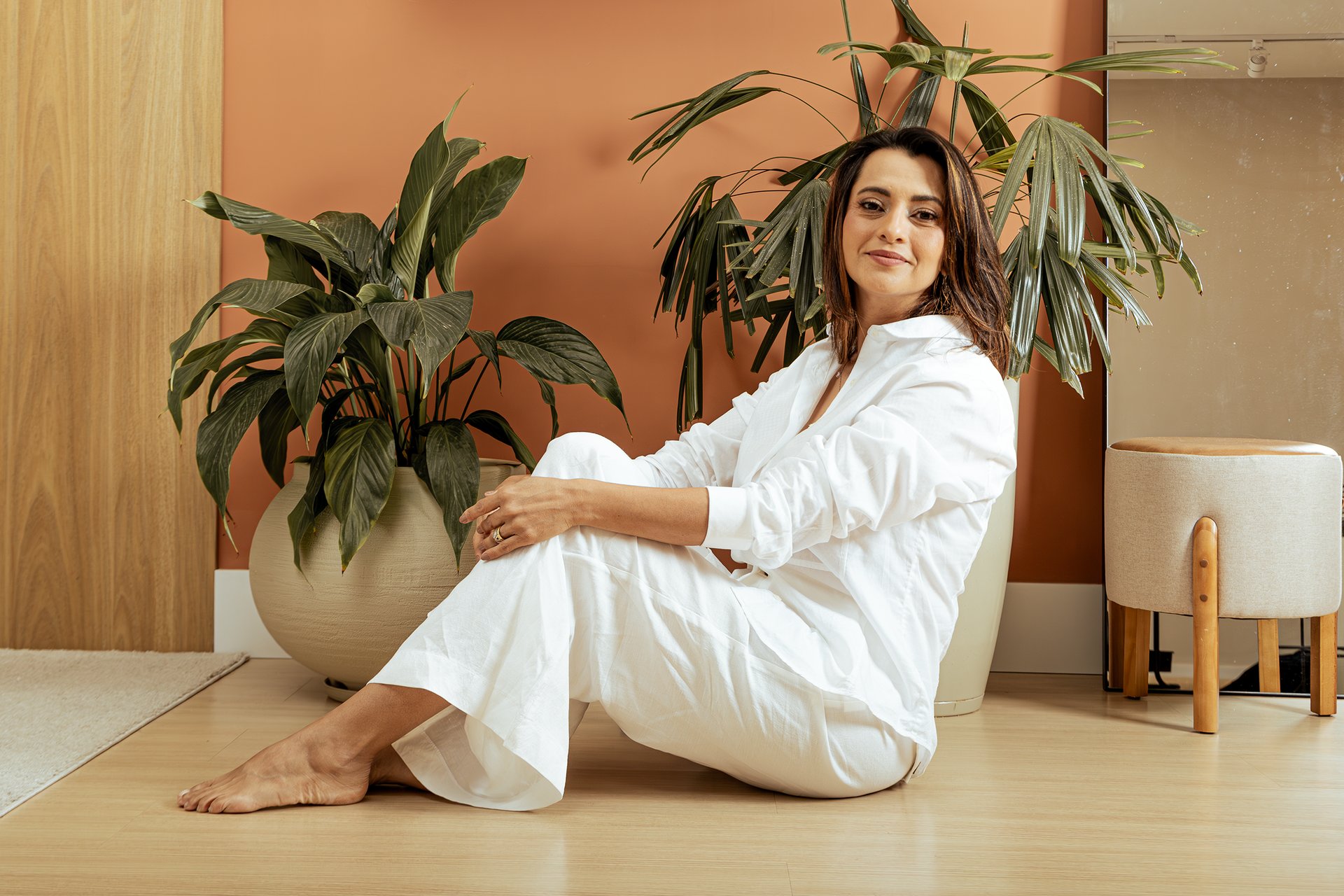 Mulher sorrindo de roupa branca, sentada no chão, uma parte da parede é de madeira freijó, outra, é laranja, há um vaso de plantas grande, e um espelho grande