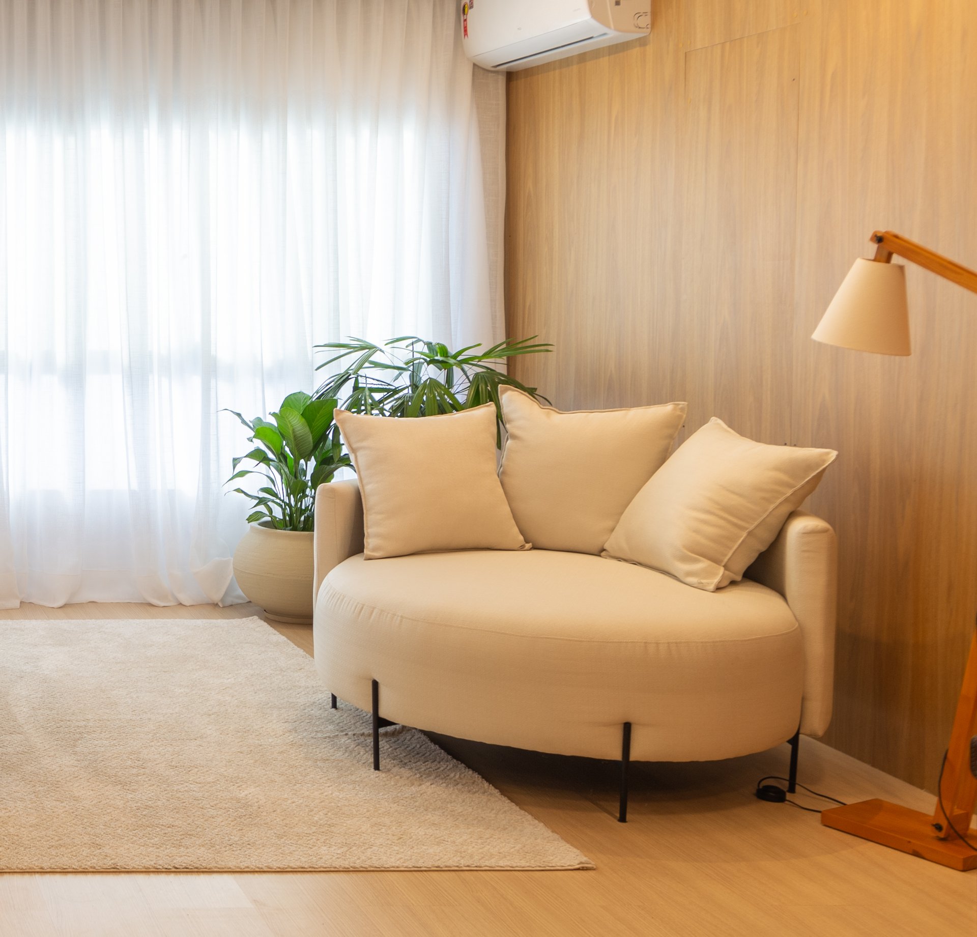 Foto de um comodo com a cortina iluminada de fundo, dois vazos de planta ao fundo, e um sofá redondo na frente, a parede é de madeira freijó.