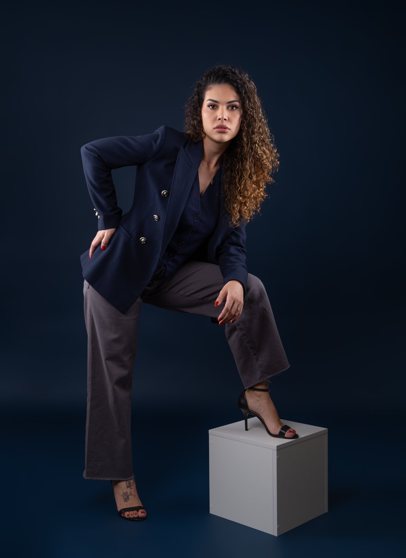 Mulher de cabelos enrolados e blazer azul, esla está de pé com uma perna apoiada em um box branco de madeira 30cm x 30cm