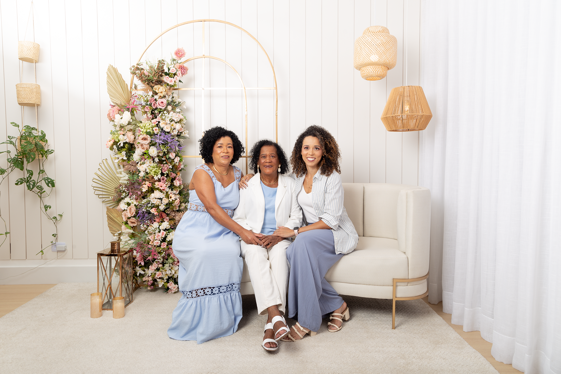 No fundo cenário do dia das mães: Sentadas no sofá da esquerda pra direita: Uma mulher (a mãe) com vestido longo azul, uma mulher mais velha com paletó branco, calça branca e camisa azul (a avó), e uma mulher mais nova com uma blusinha branca e uma camisa listrada azul por cima, e calça azul, as três mulheres são negras de cabelos crespos