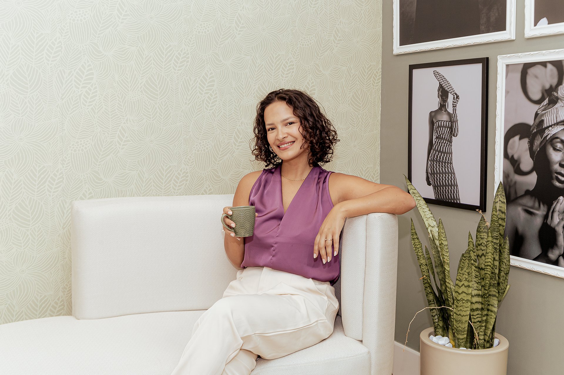Um canto com um sofá e vários quadros na parede verde musgo, Mulher branca de cabelos enrolados na altura do pescoço sentada no sofá ela está sorrindo vestida de blusinha roxa e calça bege, seu braço esquerdo está apoiado nos braços do sofá, o direito segurando uma xícara de café