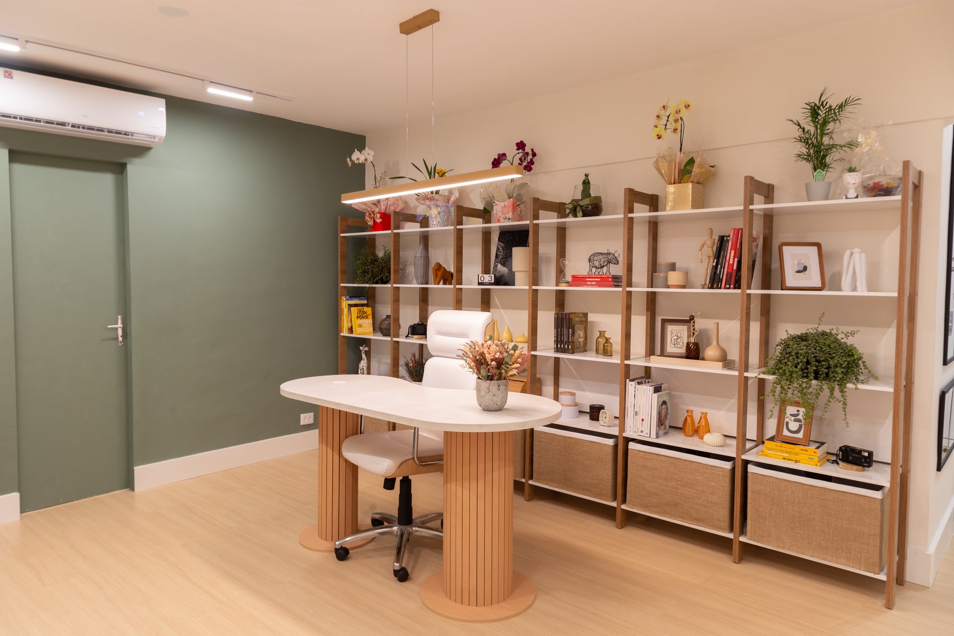 O escritório, uma parede verde musgo de fundo com quadros, no centro a mesa, e uma estante na direita