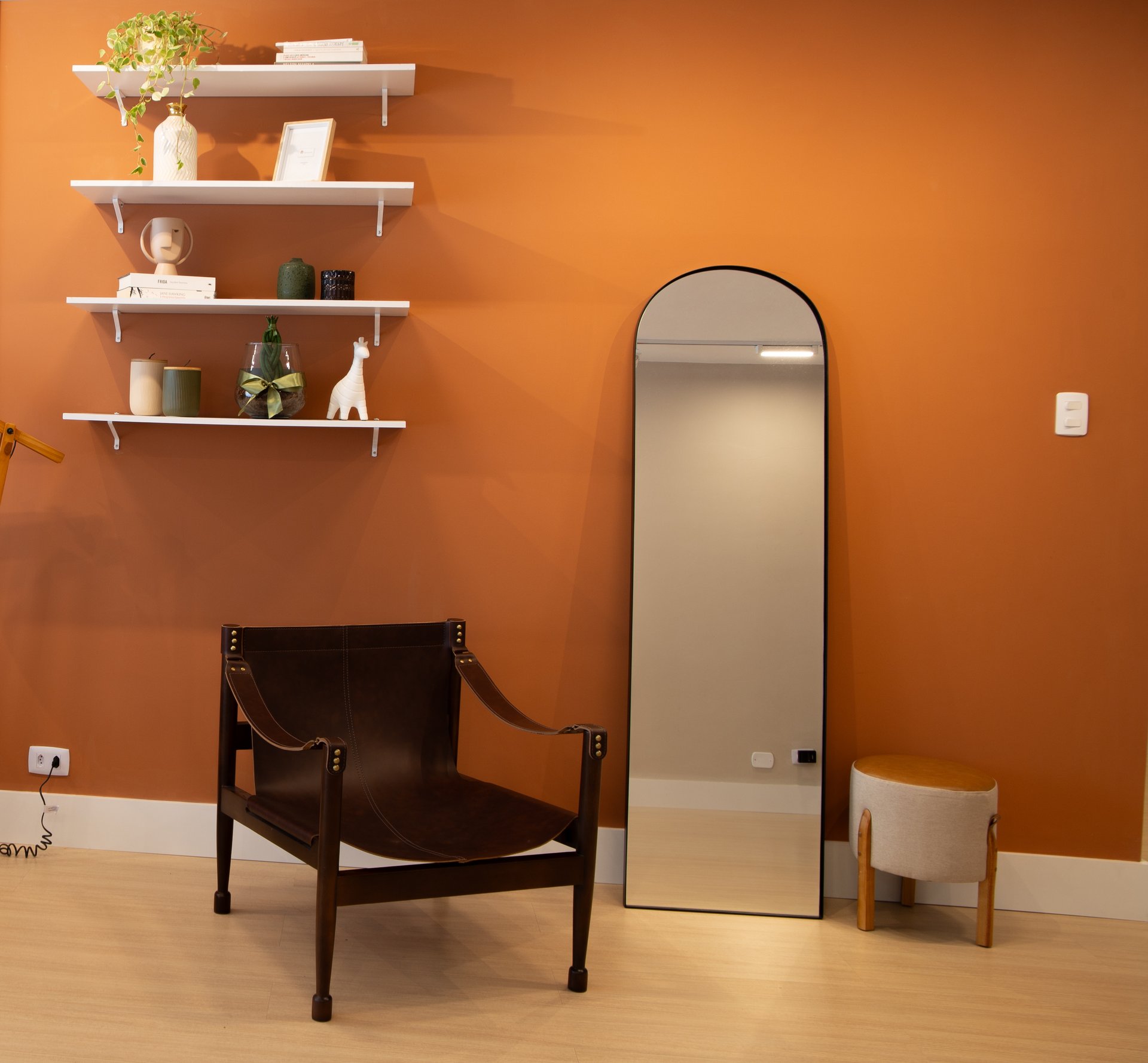 Uma parede laranja com 4 estantes brancas suspensas, uma poltrona, um espelho grande e um puff