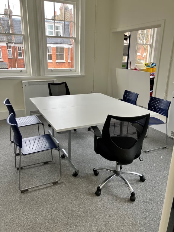 Image of meeting room one. The room has grey carpets and white walls. There are two windows shown in the image. In the centre of the room, there is a white table surrounded by six black chairs.