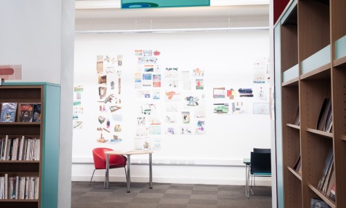 Pictures on the wall in the Pop-up Gallery behind a round table, a red and a black chair. A light blu bookshelf at the entrance of the room.
