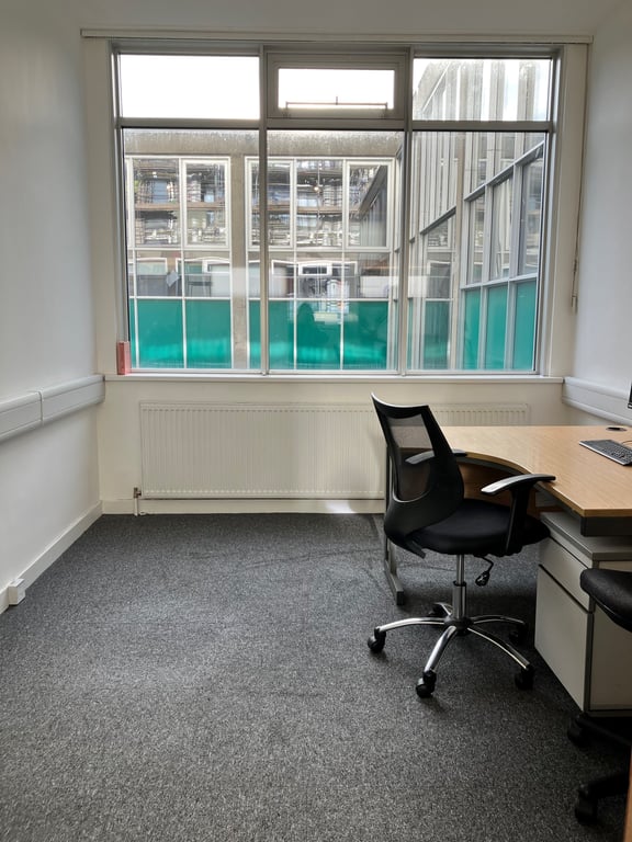 Image of meeting room three. The walls are white. The carpet is grey. The back wall of the room has a triple window. To the right in the image there is a desk with two chairs.