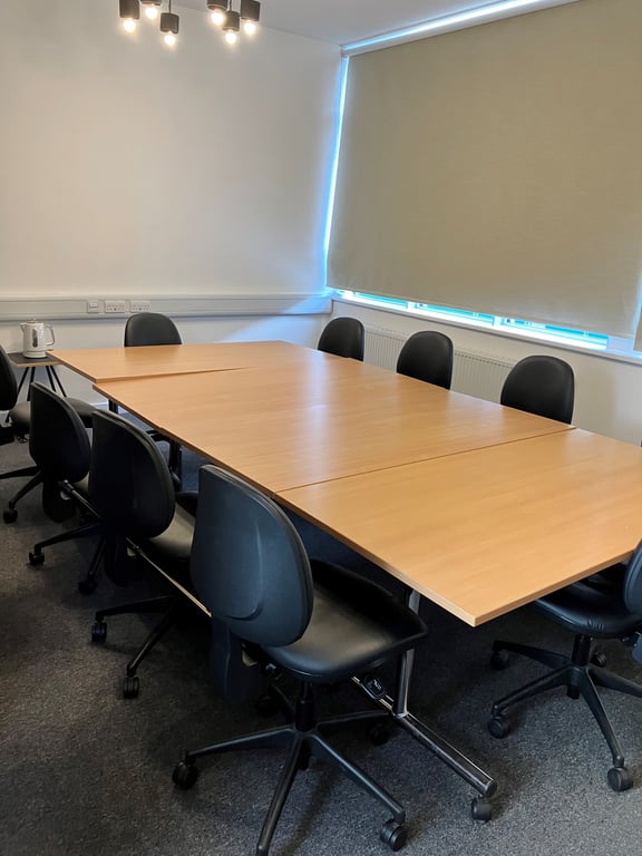 Image of meeting room five. It has white walls and grey carpets. The image shows a brown table surrounded by eight black chairs. 
