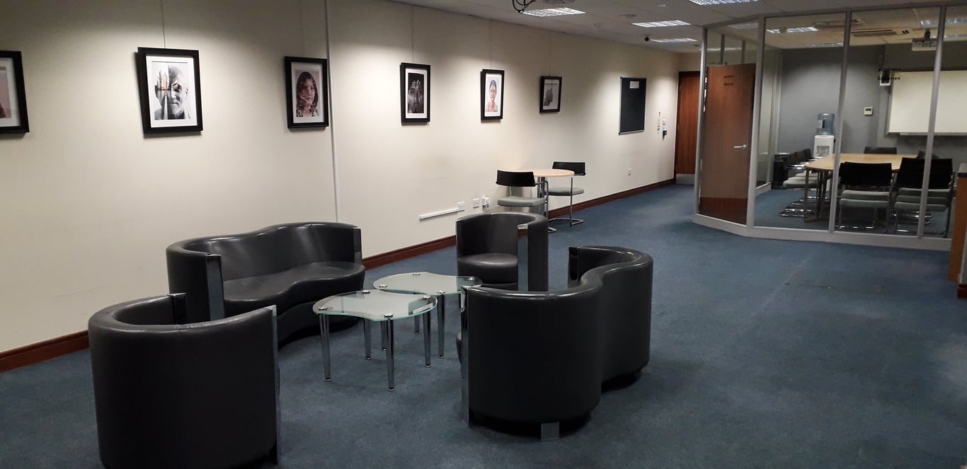This is an image of the business lounge main area. The floors are dark blue carpet and the walls are white. On the walls there is a row of black-framed portraits. There is a small glass coffee table surrounded by black arm chairs, two single chairs and two double chairs. Behind this, there is a small round table with two chairs. On the right hand side of the image there is the glass room.