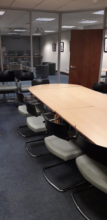 This is an image of the business lounge glass room. It shows a long brown table surrounded by grey chairs. The walls are made of glass and the door is dark brown. The floor has a dark blue carpet.