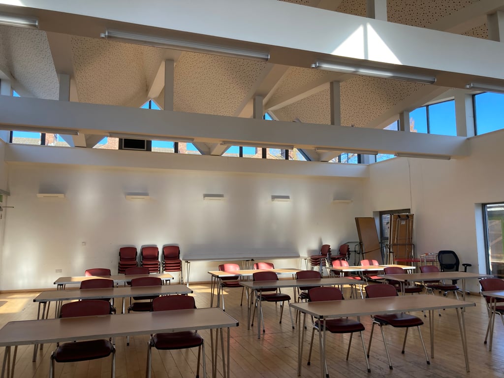 Image of the hall with desks set up. There are six brown tables set up. Each table has two red chairs behind it. The floor is brown and laminate. The walls are white. 