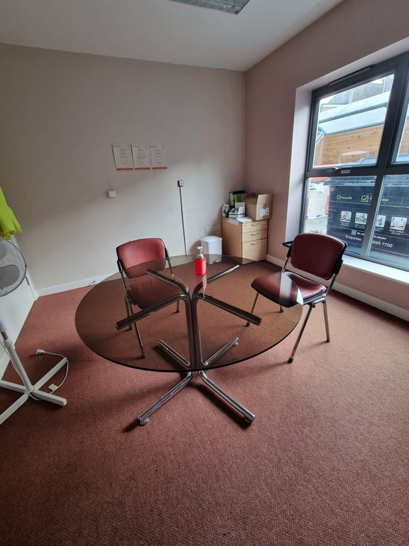 This is an image of the office room. The walls are white and the carpet is dark pink / red. The wall on the right side of the picture shows a large window. I the middle of the room there is a black, round glass table with two red chairs. There is a standing fan and white board on the left hand side of the image.