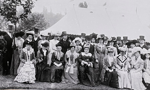 Black and white old days picture, many men and women in fancy victorian attires and hats.