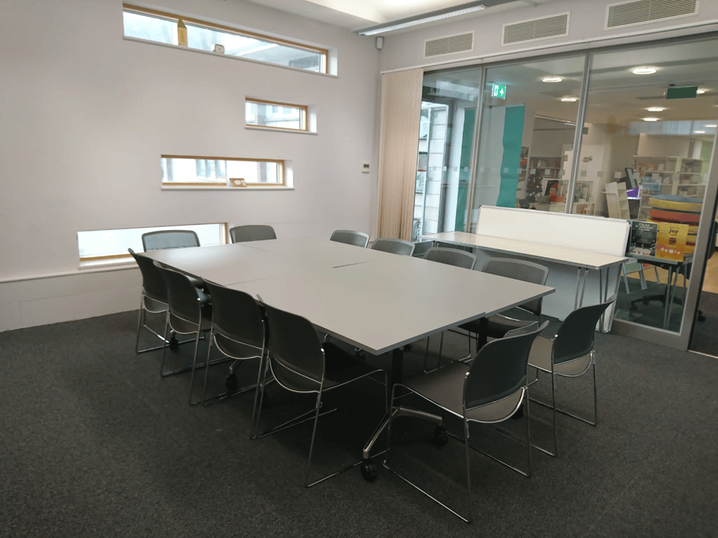 Image of Coombes Croft meeting room. The image shows a grey table surrounded by grey chairs. One of the room's walls is a glass wall. The other wall displayed is grey with four narrow windows. The floor has a dark grey carpet.