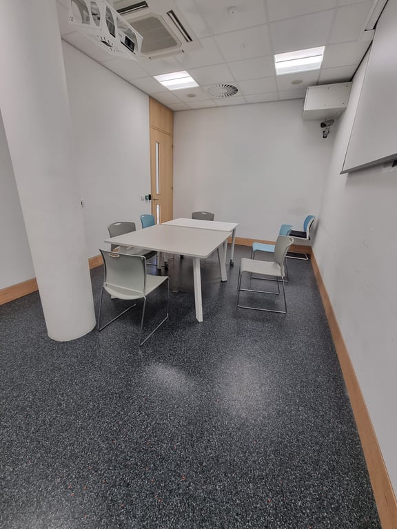 Image of meeting room one. It has white walls and a dark grey floor. In the centre of there are two tables pushed together surrounded by six chairs. The wall on the right hand side of the image has a projector screen on it.