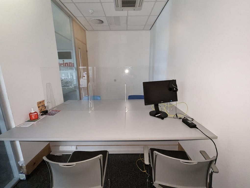 Image of meeting room three. The image shows a white table surrounded by four chairs. There is a computer on the table and a clear screen in the middle of the table. The wall on the left hand side of the image has large glass panels.