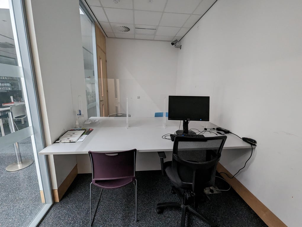 Image of meeting room three. The image shows a white table surrounded by four chairs. There is a computer on the table and a clear screen in the middle of the table. The wall on the left hand side of the image has large glass panels.