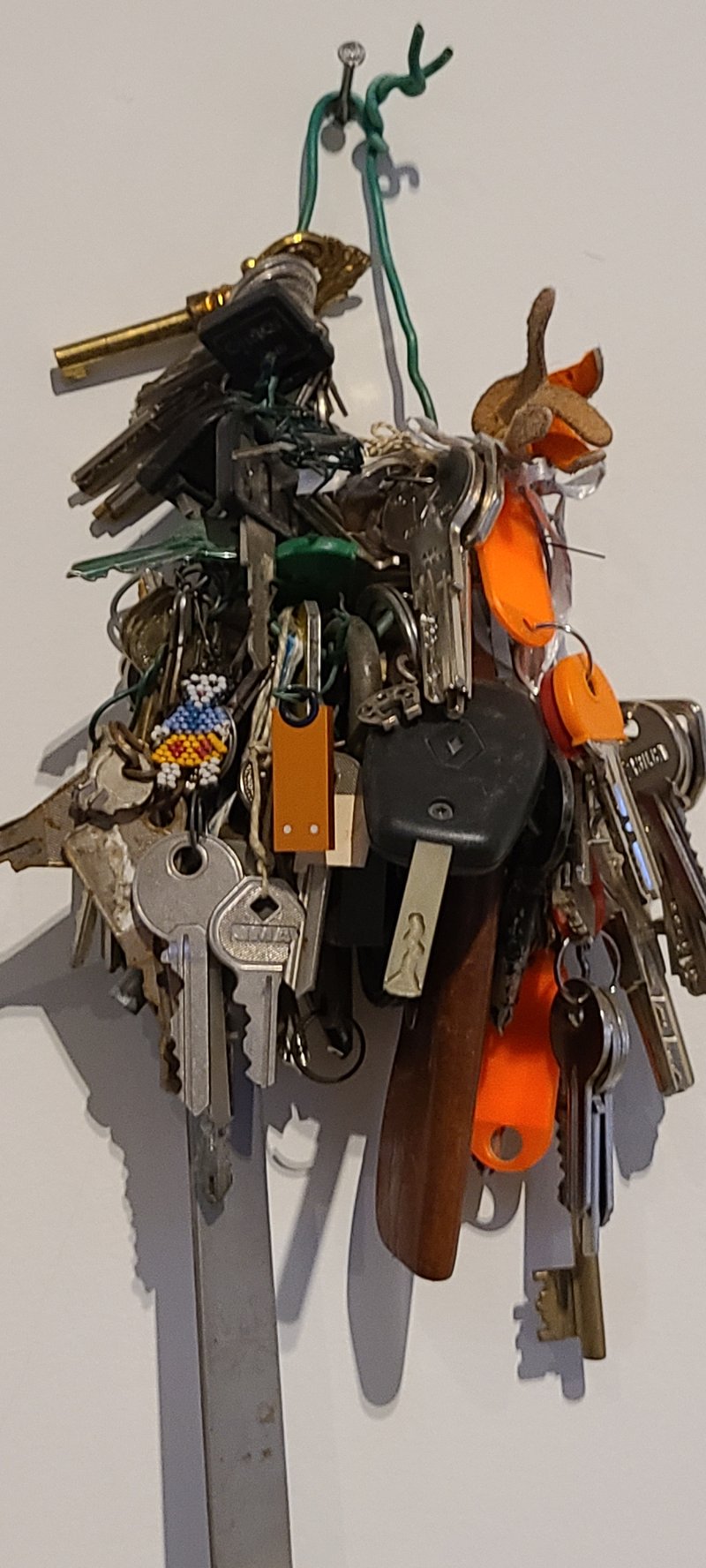 A tangled bunch of keys, tags and keychains hanging from a green wire on a nail against a white wall.
