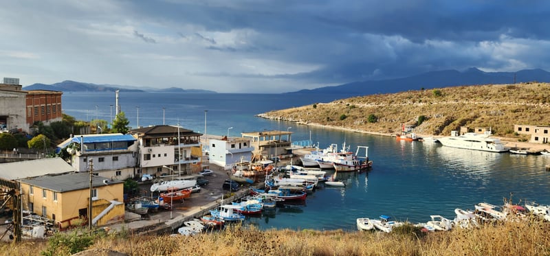 Sarandë harbor under heavy clouds: fishing boats and a white yacht moored in blue water, low buildings on the shore and mountains across the sea.