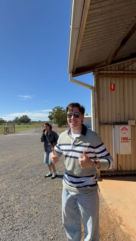 Teammate giving two thumbs up beside a farm building on gravel