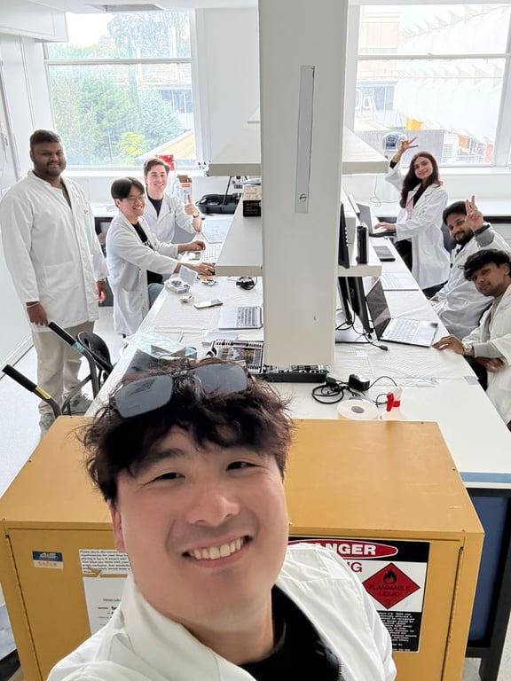 7 teammates in white coats smiling for a selfie in a lab space