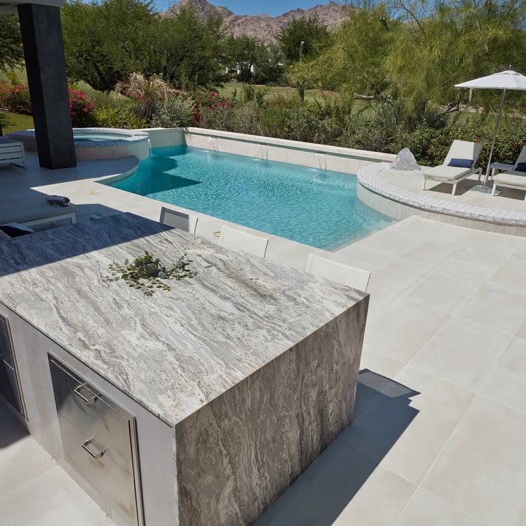 Fantasy Brown Polished marble on counters, Reside Beige 24 x 48 R11 Finish porcelain on patio floor, Reside Beige Straight Stack 1 x 12 Straight Stack porcelain on waterline & Geo 2 Metro Uptown glass tile on pool accent in outdoor patio.