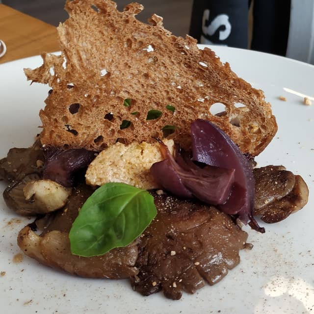 Foto do prato “Fungo Pleurotus Piastrato con Formaggino Erborinato, Cipolla Caramellata, Mandorle Tostate, Chips di Pane e Fondo Bruno Vegetale”