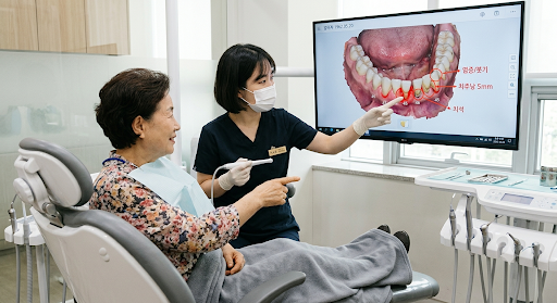 치과 진료실에서 치과의료진이 모니터 화면의 치아 및 잇몸 3D 스캔 영상을 가리키며 고령의 환자에게 치주 질환 상태를 설명하고 있는 모습