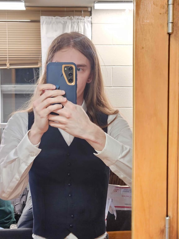 A young man with blue eyes and long blond hair stares at the camera. He is wearing a close-fitted blue vest over a black turtleneck. His hands are shoved into the pockets of a pair of pinstripe trousers.