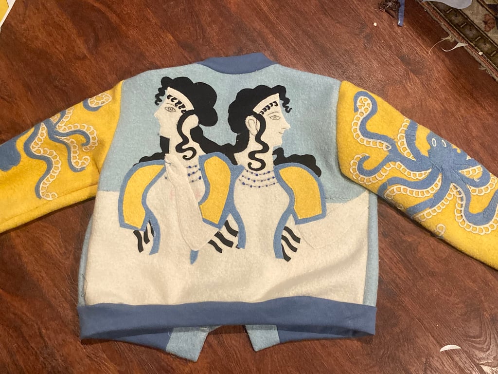 The back view of a blue and yellow jacket, with the Ladies in Blue fresco appliqued on the back