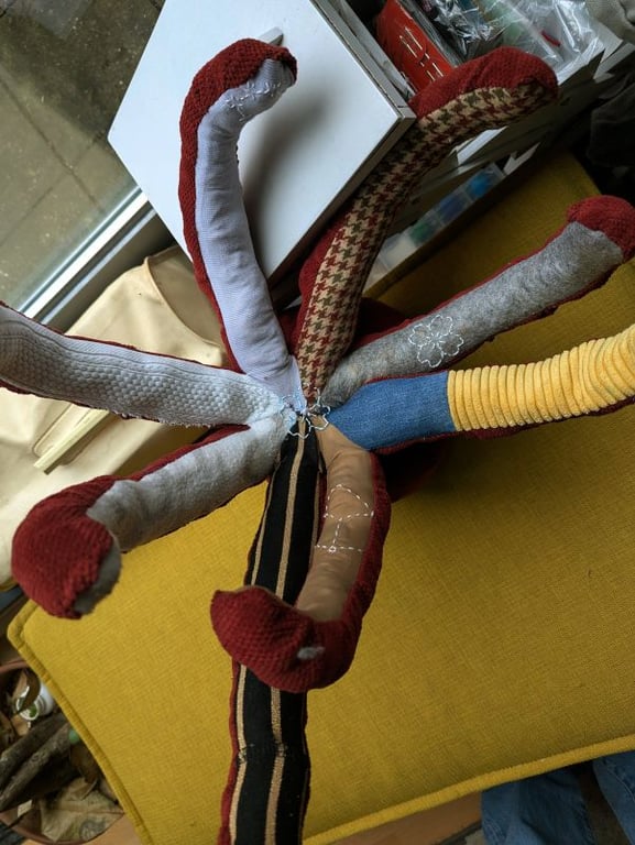 'A close-up view of the underside of one octopus plushie, showing the central point where eight tentacles meet. Each tentacle features a unique fabric lining, including grey waffle knit, brown houndstooth, grey felt with a white stitched flower, yellow corduroy, blue denim, and tan fabric with a stitched constellation pattern. The stitching at the center is visible in blue thread.'