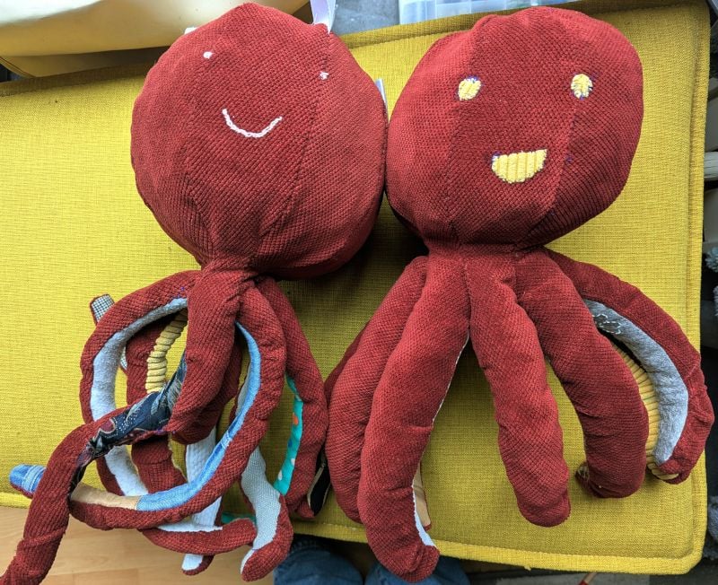 'A top-down view of two handmade octopus plushies lying side-by-side on a yellow textured surface. Both are made from a deep red, ribbed fabric. The octopus on the left has a simple white stitched smile and tiny dot eyes, with its tentacles showing various colorful patterned linings like blue denim and floral prints. The octopus on the right has larger, yellow stitched eyes and a wide, rectangular yellow stitched mouth.'