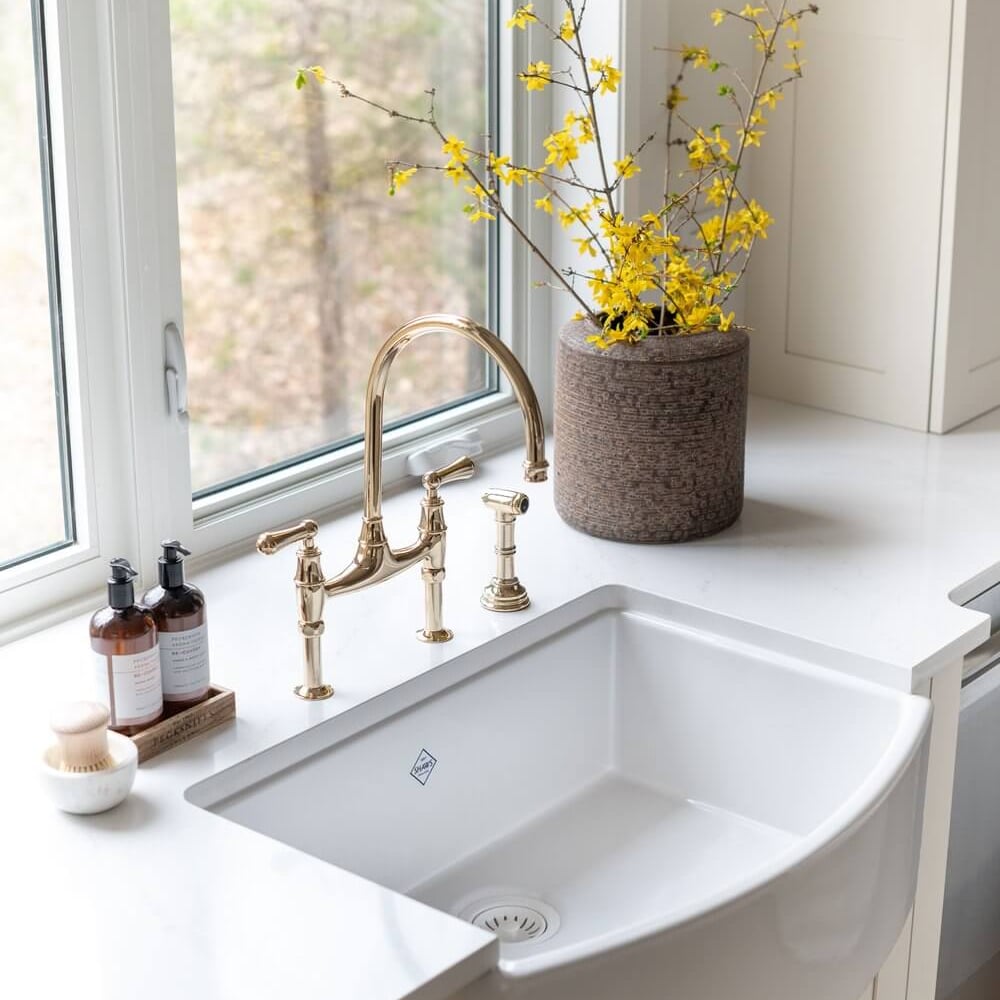 White farmhouse sink with a gold faucet and a vase of yellow flowers on a countertop.