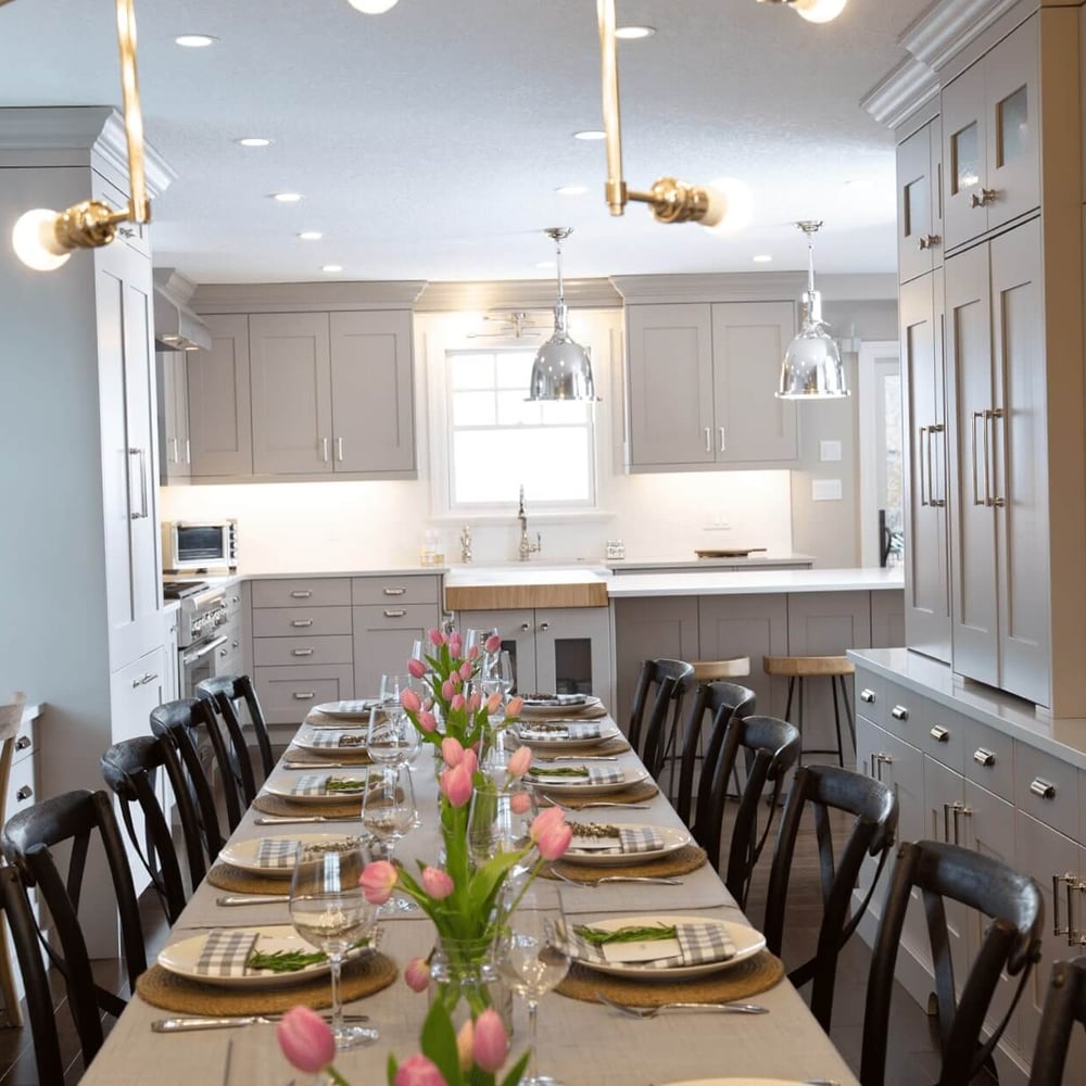 Project_Kitchens_Eastbridge10 Modern kitchen with a long dining table set with plates and glasses, featuring light gray cabinetry and floral centerpieces.