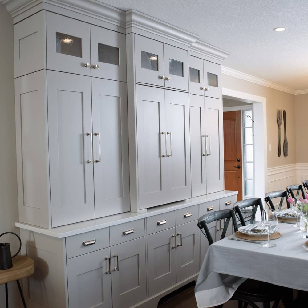 Project_Kitchens_Eastbridge11 Gray kitchen cabinetry with glass panel accents and a long dining table set with flowers and tableware in a well-lit room.